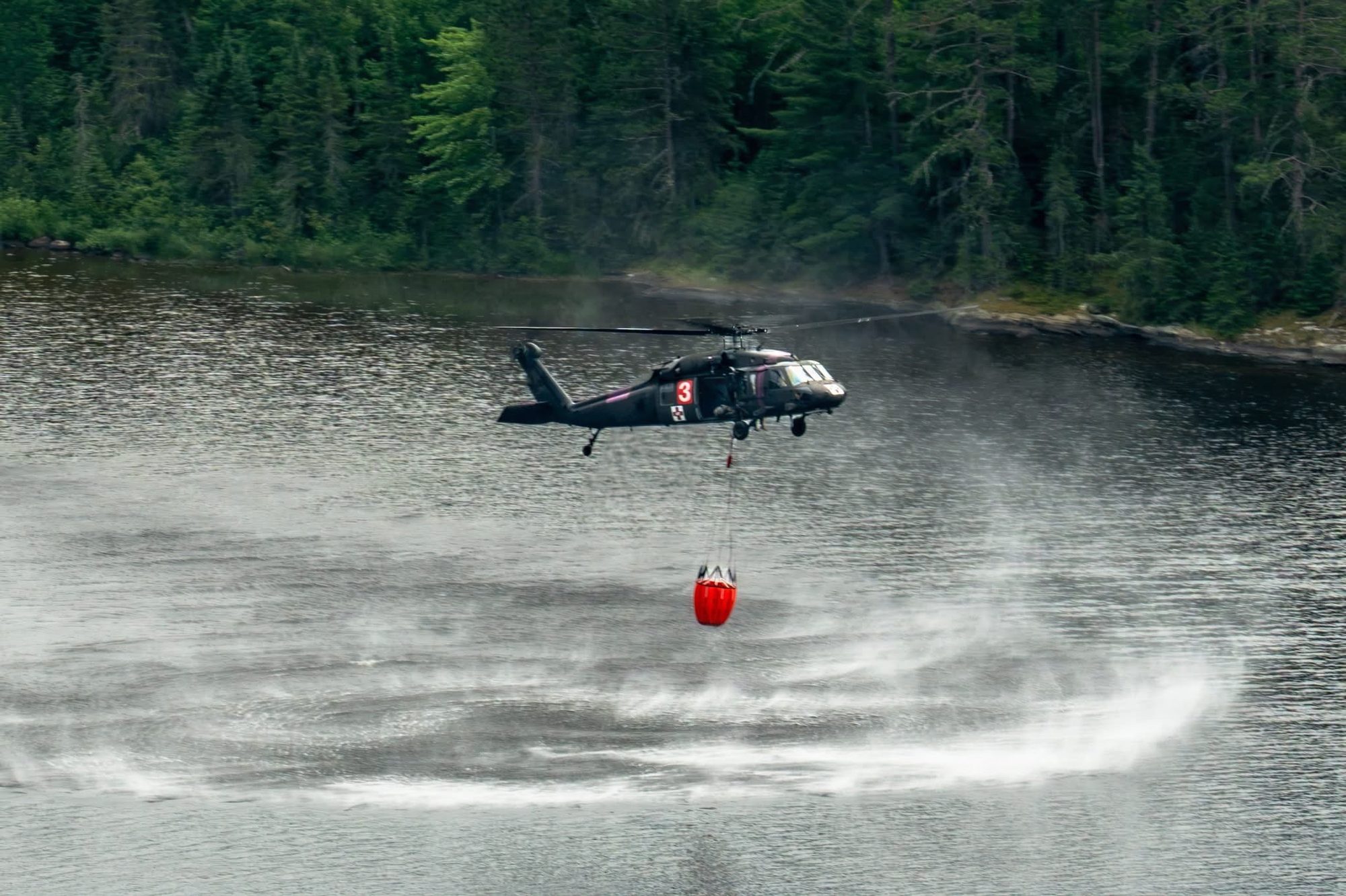 A Black Hawk helicopter hovers low over a lake, refilling a water bucket suspended beneath it as rotor wash ripples the surface.