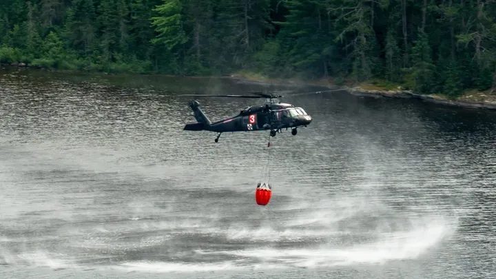 A Black Hawk helicopter with a red water bucket hovers over a lake, spray fanning out across the water beneath it.